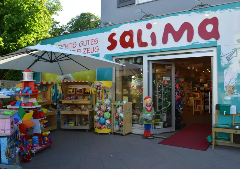 Der Eingang von Salima Kinderschätze in der Waitzstraße: türkise Fassade mit dem Schriftzug Salima, Spielzeug vor der Tür, großer Sonnenschirm